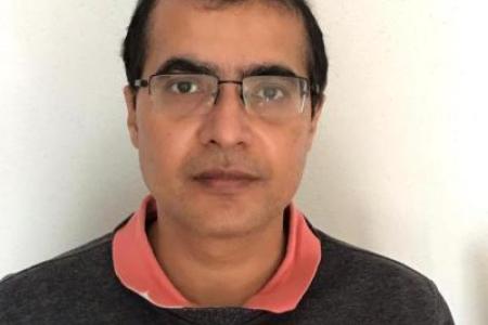 Dr. Mishra sports a pink collar over a grey sweater. Close cropped hair and bespectacled, Dr. Mishra stares into the camera with a contemplative expression. 