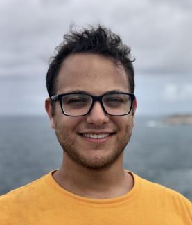 Dr. Coleman smiles at the camera in front of the ocean. He sports a yellow shirt and square black glasses. 