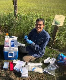 Dr. Jose Rolando sits in front of an array of sampling equipment. He kneels on the ground smiling at the camera. He is wearing field clothes including tan cargo pants and a navy button up with glove covered hands. 