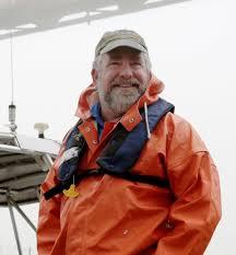 Dr. Sam Bentley smiles at the camera in a orange slicker with emergency life jacket and tan baseball cap