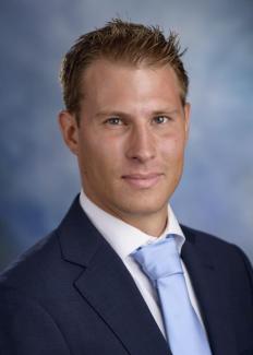 Dr. Stefano Carattini stares at the camera in a navy suit coat, white collar shirt, and light blue tie. His hair is close cropped and brown. 