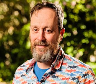 Dr. Christopher Romsos has a full beard and messy short hair. He is wearing a blue undershirt and a multi-color shark button up