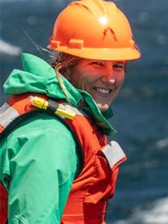 Dr. Kelly Sutherland looks over her shoulder smiling in a green jacket, orange life vest, and hard hat. 