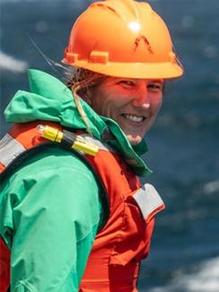 Dr. Kelly Sutherland looks over her shoulder smiling in a green jacket, orange life vest, and hard hat.