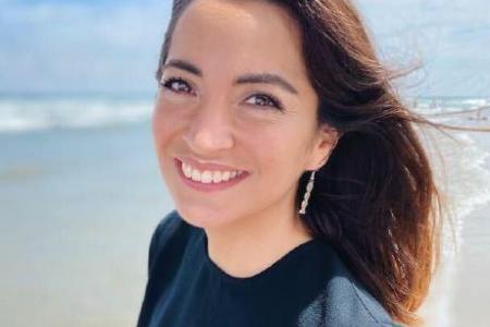 Dr. Valerie De Anda smiles into the camera on the beach with dark red-brown hair blowing in the wind