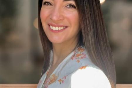 Dr. Valerie De Anda stands sideways with her head turned towards the camera smiling. Her dark brown hair falls behind her back against her white button up. 