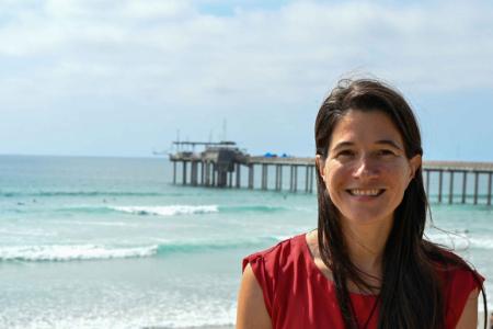 Dr. Moira Decima smiles at the camera in a shoulderless red shirt backed by the ocean and a pier. 