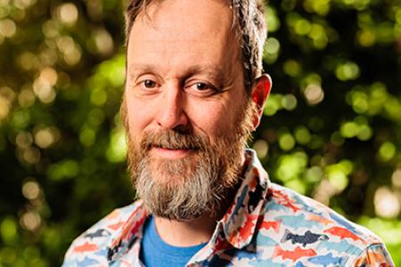 Dr. Christopher Romsos has a full beard and messy short hair. He is wearing a blue undershirt and a multi-color shark button up