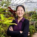 Dr. Chung smiles confidently, arms crossed, and hair pulled back. She is surrounded by greenery and stands out wearing a black cardigan and purple collared button up. 