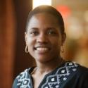 Dr Erica Holloman-Hill smiles into the camera in a black v-neck embroidered in white. Her hair is a box short natural cut and gold hoops frame her face.