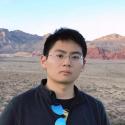 Dr. Gengchen Mai stands in from of a rock expanse wearing a black jacket, glasses with metal frames, and sunglasses on his jacket collar. He stares into the camera without smiling. 