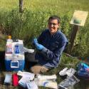 Dr. Jose Rolando sits in front of an array of sampling equipment. He kneels on the ground smiling at the camera. He is wearing field clothes including tan cargo pants and a navy button up with glove covered hands. 