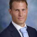 Dr. Stefano Carattini stares at the camera in a navy suit coat, white collar shirt, and light blue tie. His hair is close cropped and brown. 