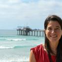 Dr. Moira Decima smiles at the camera in a shoulderless red shirt backed by the ocean and a pier. 