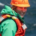 Dr. Kelly Sutherland looks over her shoulder smiling in a green jacket, orange life vest, and hard hat. 